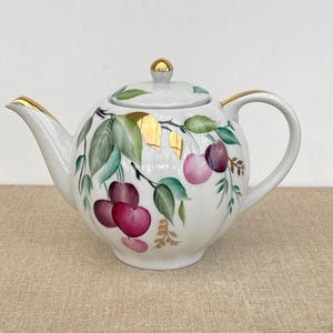 May include: White ceramic teapot with a gold-trimmed spout, handle, and lid. The teapot is decorated with a hand-painted design of cherries, green leaves, and gold accents. The cherries are shades of red and purple.