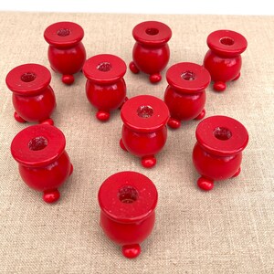 Puede incluir: Un conjunto de diez pequeños portavelas de color rojo brillante. Cada portavelas presenta un cuerpo redondeado y bulboso con una parte superior plana y circular que contiene un orificio central para una vela, apoyado sobre tres pies pequeños.