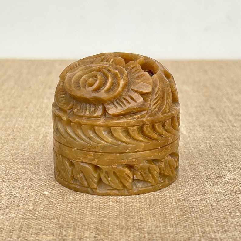 Puede incluir: Una peque&ntilde;a caja redonda de piedra tallada con un dise&ntilde;o floral. La caja est&aacute; hecha de piedra marr&oacute;n claro y tiene una tapa decorada con una rosa.