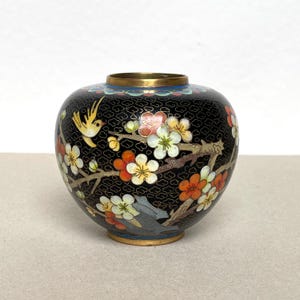 Pode incluir: Um pequeno vaso redondo preto com um desenho floral e de pássaros. O vaso apresenta uma borda e base douradas, com flores coloridas e um pássaro amarelo. As flores são brancas, laranja e rosa.