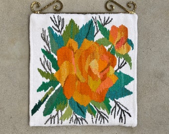 Op de afbeelding: Een wit wandkleed met een bloemenmotief met een grote oranje roos met roze accenten en groene bladeren. Het wandkleed is omlijst met een gouden metalen staaf.