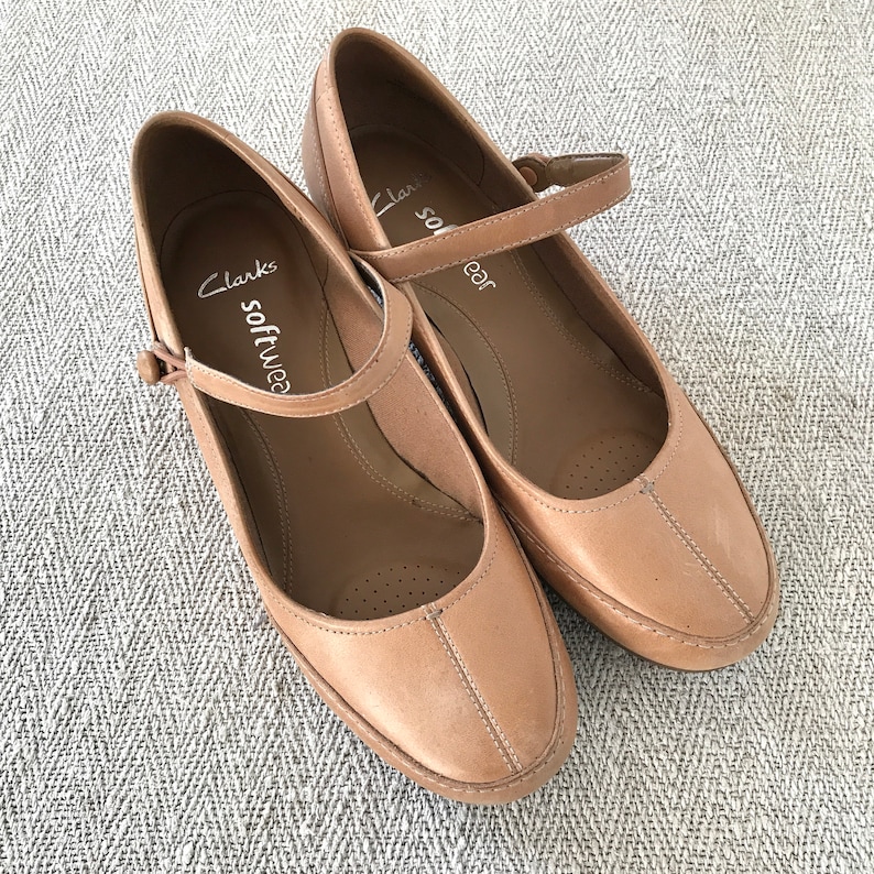 K&ouml;nnte beinhalten: Ein Paar hellbraune Clarks Softwear Mary Jane Schuhe. Die Schuhe haben eine runde Spitze, einen Riemen mit Schnalle und Ziern&auml;hte. Sie liegen auf einer hellgrauen, gemusterten Oberfl&auml;che. Klassischer Schuhstil.