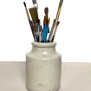 Könnte beinhalten: Eine Sammlung von Pinseln verschiedener Größen und Formen in einem beigefarbenen Keramiktopf. Die Pinsel haben Holzgriffe und Borsten in Braun-, Schwarz- und Blautönen. Der Topf hat eine runde Form und der Text "DAMOY PARIS" ist sichtbar.