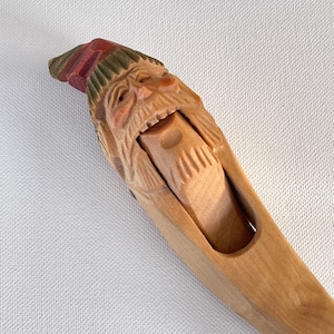 May include: A wooden nutcracker carved to resemble an old man with a long beard and a hat. The hat is painted red and green. The nutcracker's mouth is open, showing the mechanism for cracking nuts.