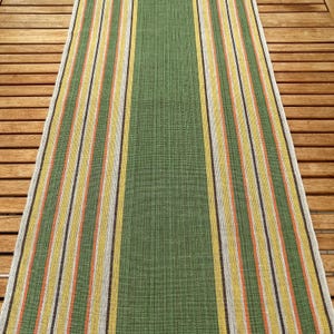 May include: A green and striped runner rug with vertical stripes in yellow, orange, white, and brown. The rug has a solid green center and is likely made of woven material. The rug is placed on a wooden surface.