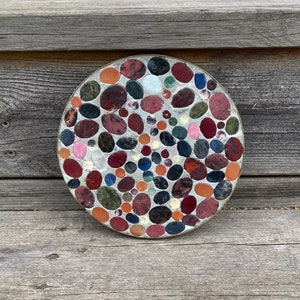 Peut inclure: Un dessous de plat rond et décoratif composé d'une mosaïque de petites pierres colorées. Les pierres sont disposées de manière aléatoire et sont principalement de couleur rouge, marron et noire.