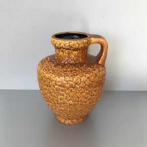 Puede incluir: Un jarrón de cerámica marrón con una superficie texturizada y un asa. El jarrón tiene un cuerpo redondeado y un cuello estrecho.