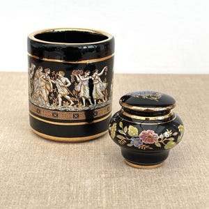 Può includere: Un contenitore in ceramica nera e un piccolo barattolo con coperchio. Il contenitore ha un bordo dorato e un fregio raffigurante figure in abiti classici. Il barattolo è decorato con motivi floreali e dettagli dorati.