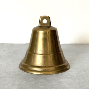 Puede incluir: Una pequeña campana de latón de estilo antiguo, con una base redondeada y un lazo en la parte superior para colgarla. La campana tiene una superficie lisa y pulida con una ligera pátina, lo que le da un aspecto vintage. La campana es de color dorado cálido.