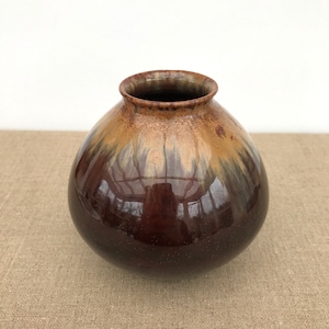May include: A small, round, brown ceramic vase with a glossy glaze. The vase has a light brown and white glaze on the top half, creating a mottled effect.