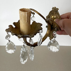 Può includere: Un'applique da parete in ottone con una piastra posteriore ornata, un braccio curvo e un portacandele centrale con un manicotto che imita la cera gocciolante. L'apparecchio è decorato con diversi pendenti in cristallo trasparente sfaccettato.
