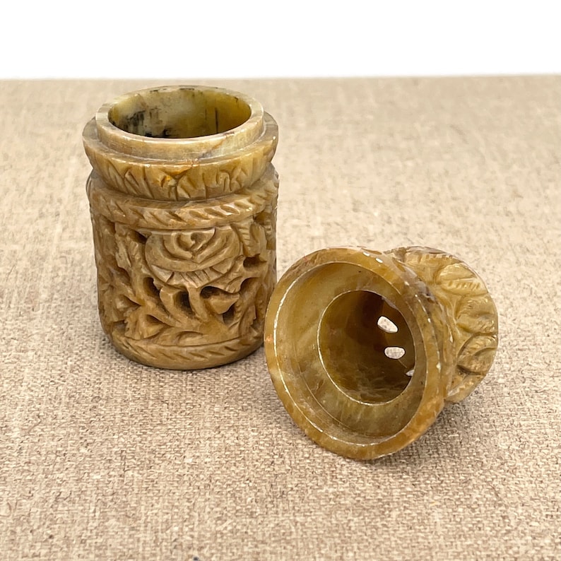 Puede incluir: Un peque&ntilde;o recipiente de piedra beige tallado con tapa. El recipiente tiene intrincados dise&ntilde;os florales y est&aacute; abierto para revelar el interior.