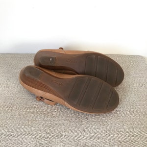 K&ouml;nnte beinhalten: Ein Paar Clarks Schuhe mit Keilabsatz in hellbrauner Farbe. Die Sohlen sind dunkelbraun mit einem strukturierten Muster. Die Schuhe haben einen Riemen mit Schnallenverschluss. Gr&ouml;&szlig;e 6.