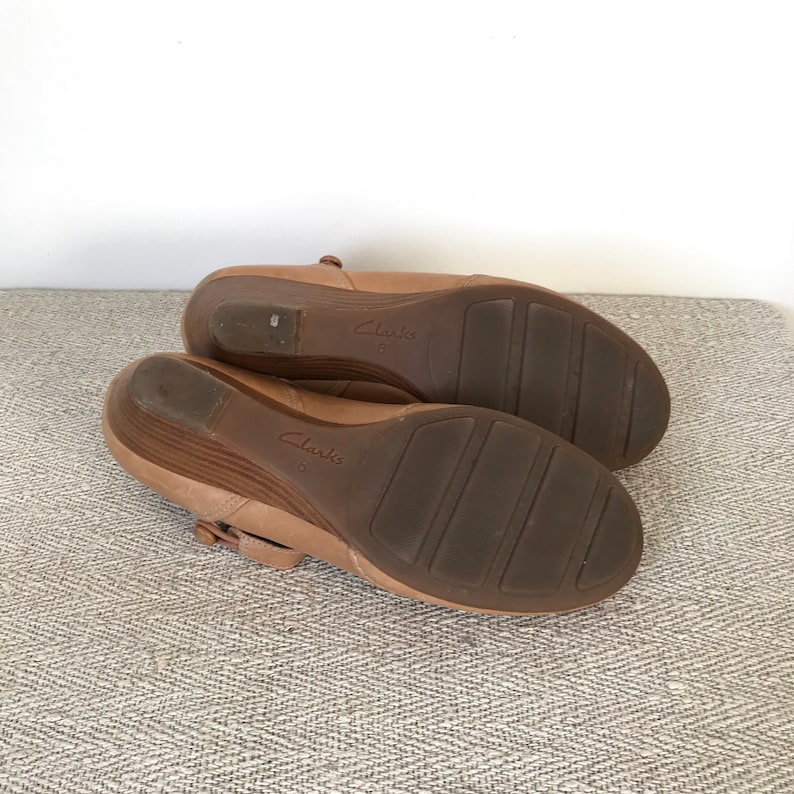 K&ouml;nnte beinhalten: Ein Paar Clarks Schuhe mit Keilabsatz in hellbrauner Farbe. Die Sohlen sind dunkelbraun mit einem strukturierten Muster. Die Schuhe haben einen Riemen mit Schnallenverschluss. Gr&ouml;&szlig;e 6.