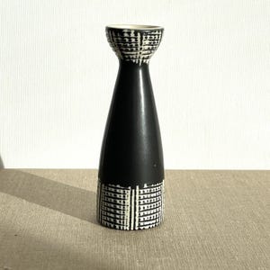 Puede incluir: Un candelero de cerámica en blanco y negro. La base y la parte superior del candelero tienen un patrón geométrico blanco y negro. La sección media es negra. El candelero está sobre una superficie neutra.