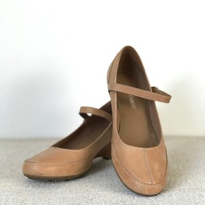 K&ouml;nnte beinhalten: Ein Paar hellbraune Mary Jane-Schuhe aus Leder mit kleinem Keilabsatz. Die Schuhe haben einen Riemen &uuml;ber dem Fu&szlig; und eine abgerundete Spitze. Die Schuhe werden auf einer strukturierten Oberfl&auml;che pr&auml;sentiert.