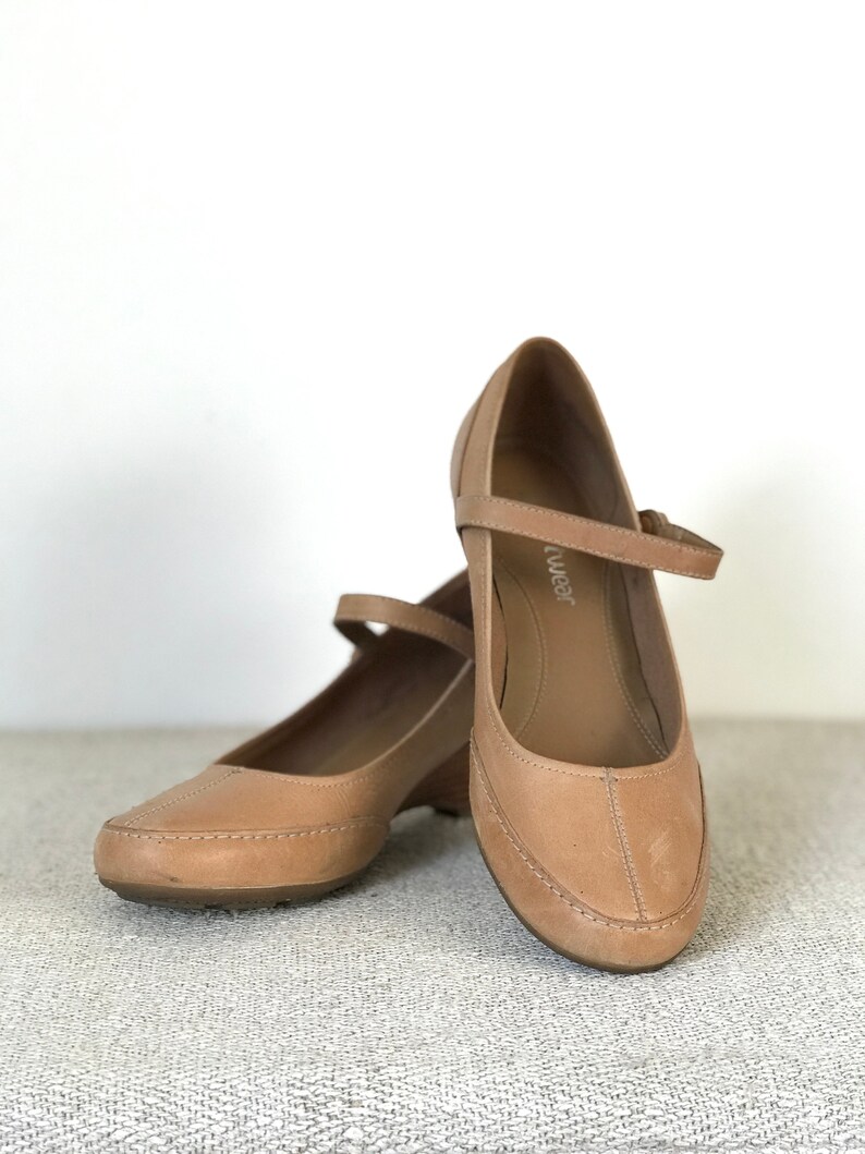 K&ouml;nnte beinhalten: Ein Paar hellbraune Mary Jane-Schuhe aus Leder mit kleinem Keilabsatz. Die Schuhe haben einen Riemen &uuml;ber dem Fu&szlig; und eine abgerundete Spitze. Die Schuhe werden auf einer strukturierten Oberfl&auml;che pr&auml;sentiert.