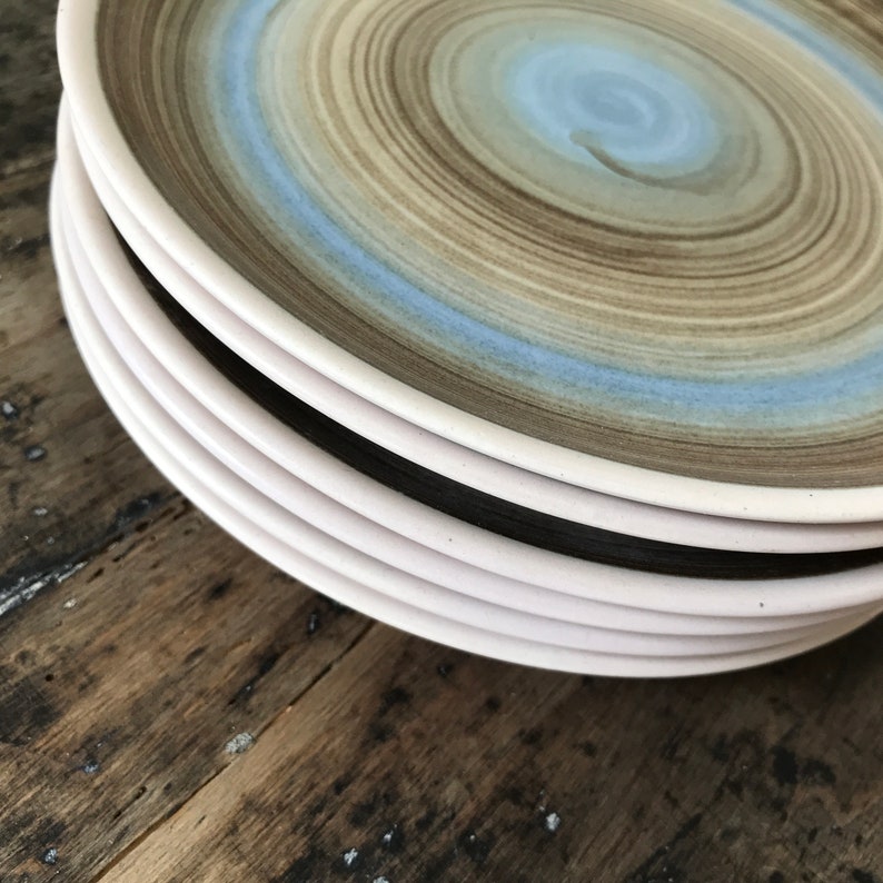 May include: A stack of ceramic dinner plates with a brown and blue swirl design. The plates have a white rim and are stacked on a wooden surface.