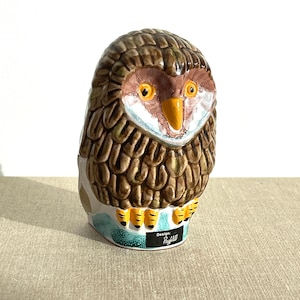 Puede incluir: Figura de búho de cerámica con detalles marrones, blancos y naranjas. El búho tiene un patrón de plumas detallado, un pico amarillo y ojos amarillos. La base del búho es turquesa con el texto "Design Paghild".