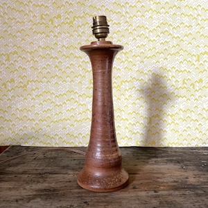 Puede incluir: Una lámpara de mesa de cerámica marrón con un diseño sencillo. La lámpara tiene una base alta y cilíndrica y un porta-pantalla redondo. La lámpara está sobre una superficie de madera.