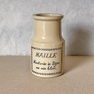 Puede incluir: Un tarro de cerámica beige con forma cilíndrica y una parte superior ligeramente ensanchada. El tarro presenta una etiqueta azul con la palabra "MAILLE" y el texto "Moutarde de Dijon au vin blanc". La etiqueta está enmarcada con un borde decorativo azul.