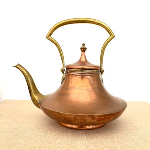 Puede incluir: Una tetera de cobre con asa y tapa de latón. La tetera tiene un cuerpo redondeado y un pico en el lateral.