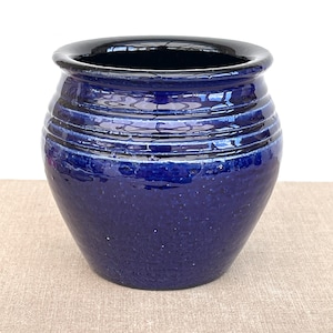 May include: A deep cobalt blue ceramic pot with a glossy, reflective finish. The vessel features a rounded, bulbous body shape with a series of horizontal, incised grooves encircling the upper section just below the wide, smooth, black-rimmed opening.
