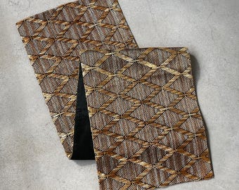 May include: A woven table runner with a geometric diamond pattern in shades of brown, tan, and cream. The runner is folded, revealing a black fabric on the underside. The texture appears to be made of natural fibers.