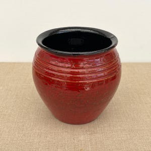 May include: A red ceramic pot with a black rim. The pot has a textured surface and horizontal ridges around the body. The interior is black. The pot is displayed on a neutral-coloured surface.