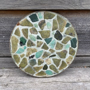 Puede incluir: Un posavasos redondo de mosaico verde y blanco con un borde de latón. El posavasos está hecho de pequeñas piezas irregulares de piedra.