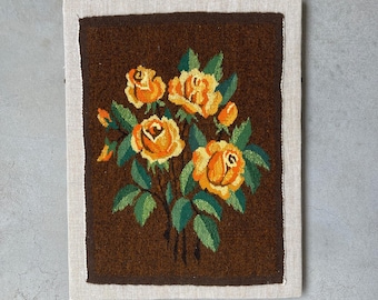 Op de afbeelding: Een ingelijste tapisserie met een boeket gele en oranje rozen met groene bladeren tegen een donkerbruine achtergrond. Het kunstwerk is omlijst met een lichtbeige rand.