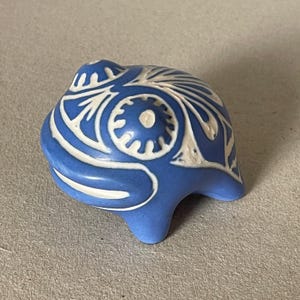 Może przedstawiać: Niebieski ceramiczny przedmiot dekoracyjny z białymi wzorami kwiatowymi. Obiekt ma zaokrągloną górę i trzy małe nóżki. Biały wzór jest kontrastującym elementem na niebieskim tle. Obiekt jest prawdopodobnie elementem dekoracyjnym.