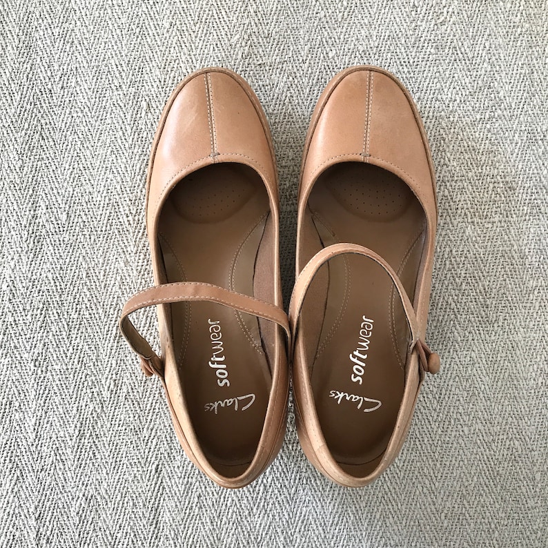 K&ouml;nnte beinhalten: Ein Paar hellbraune Mary Jane-Schuhe aus Leder mit Riemen und Schnalle. Die Schuhe haben eine runde Spitze und einen niedrigen Absatz. Der Markenname "Clarks softwear" ist auf der Innensohle sichtbar.
