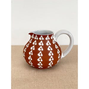 May include: A ceramic pitcher with a rounded body and a white handle. The pitcher is a deep reddish-brown color and is decorated with a repeating pattern of white, stylized heart shapes. The interior of the pitcher is white.
