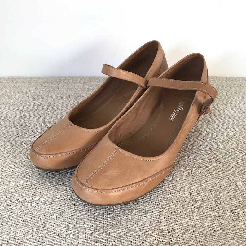 K&ouml;nnte beinhalten: Ein Paar hellbraune Mary Jane-Schuhe aus Leder mit Riemen und Schnalle. Die Schuhe haben eine runde Spitze und einen kleinen Absatz. Der Markenname "softwear" ist auf der Innensohle sichtbar.