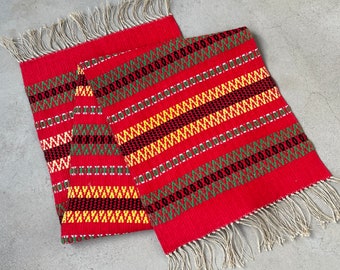 Può includere: Due tovagliette all'americana in tessuto rosso con bordi a frangia. Le tovagliette hanno un motivo geometrico a strisce nere, gialle e verdi.
