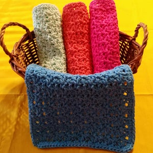 May include: A woven wicker basket holds four rolled washcloths in shades of green, red, and pink, with a blue crocheted washcloth in front. The items are on a yellow surface.