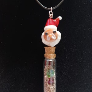 Può includere: Una collana con un flacone di vetro e un ciondolo di Babbo Natale. Il flacone è pieno di sabbia e piccole perline di vetro colorate. La collana è su un cordoncino nero.