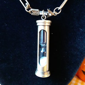 May include: A gold-toned necklace with an arrow charm and a small hourglass pendant. The hourglass is filled with white sand.