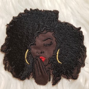May include: Embroidered patch featuring a Black woman with closed eyes, red lips, and gold hoop earrings. Her dark hair is voluminous and curly. The patch is a dark brown color with a textured, frayed edge, and is set against a white, fluffy background.
