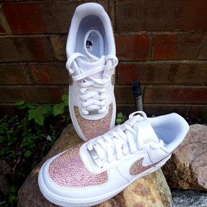 May include: White sneakers with pink and gold crystal embellishments on the toe and Nike swoosh. The shoes have white laces and a white sole. The sneakers are displayed on a rock with a brick background.