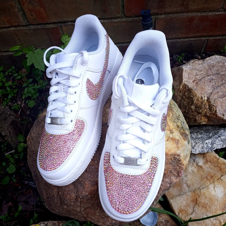 May include: Pair of white leather sneakers with pink crystal embellishments on the toe and Nike swoosh. The sneakers have white laces and a silver metal tag that says "AF1". The shoes are sitting on a rock.