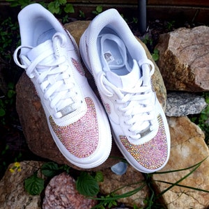 May include: Pair of white sneakers with pink and gold crystal embellishments on the toe box. The shoes have white laces, a white sole, and the iconic Nike swoosh logo on the side. The sneakers are displayed on a rock.