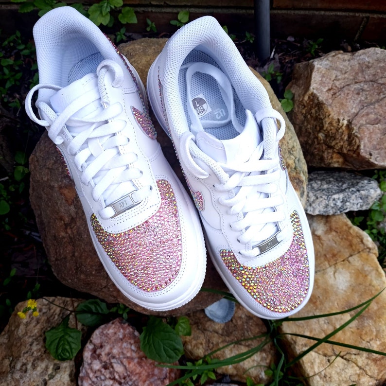 May include: Pair of white sneakers with pink and gold crystal embellishments on the toe box. The shoes have white laces, a white sole, and the iconic Nike swoosh logo on the side. The sneakers are displayed on a rock.