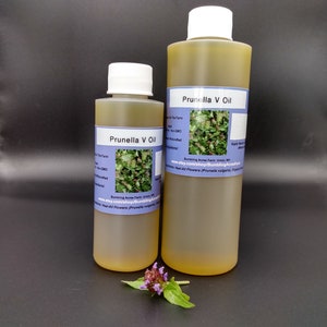 Puede incluir: Dos botellas de vidrio ámbar de aceite de Prunella V, un remedio herbal. Las botellas están etiquetadas con el nombre del producto y la dirección del sitio web de Bumbling Acres Farm. La botella más pequeña está delante de la más grande. Una pequeña flor morada está en primer plano.