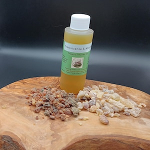 Puede incluir: Una botella de aceite orgánico infundido de incienso y mirra con una tapa blanca, junto a dos montones de resina sobre una superficie de madera. El aceite es de color ámbar y la resina es marrón claro y oscuro.