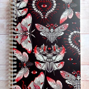 May include: Spiral-bound notebook featuring a black cover with a repeating pattern of stylized moths, insects, and eyes in red, gray, and white. The notebook has a white spiral binding and is set against a light wood grain background.