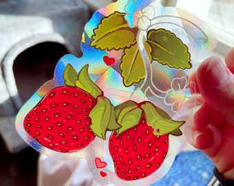 Può includere: Un adesivo olografico con due fragole rosse con foglie verdi e fiori bianchi su una vite. L'adesivo ha una finitura arcobaleno iridescente.