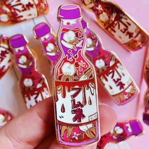 May include: A purple and gold enamel pin shaped like a Japanese ramune bottle. The bottle is filled with red and white candy, and the label says "ラムネ".