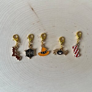 May include: Five gold-colored Halloween-themed charms with different designs: a bat, a spider web, a witch's hat, a spider, and a candy cane.